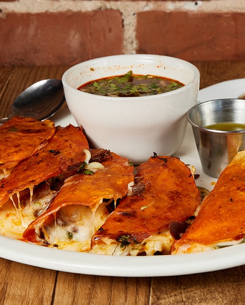 Birria quesa tacos with melting cheese and a white bowl of consome on a wooden table against exposed red brick
